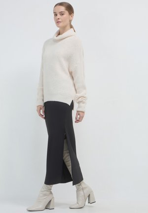 Donna che indossa un maglione a collo alto color crema, una gonna nera lunga fino alla caviglia con spacco laterale e stivaletti con tacco color crema, in piedi contro uno sfondo bianco.