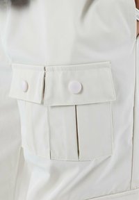 Tela de color beige claro con una textura suave, que presenta un bolsillo con tapa rematado con dos botones de presión blancos y un diseño plisado en el costado.