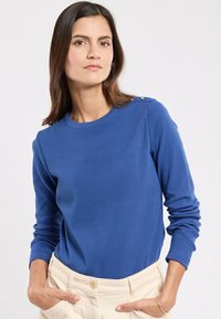 Blauwe lange mouwen trui met een geribbelde textuur en ronde halslijn, met gouden knoopdetails op de linkerschouder. Crème-kleurige broek.