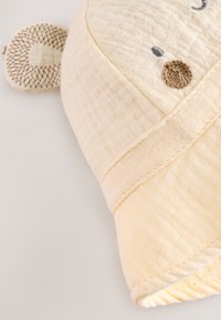 Bela in mehka muslinska otroška brisača v obliki živalske glave, z teksturirano površino, vezenimi očmi in okrasnim ušesom.