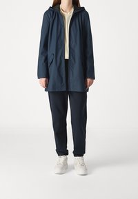 Marinblå regnjacka med huva, två framfickor och dragkedja. Bärs över en ljus topp och mörka sweatpants, tillsammans med vita sneakers.