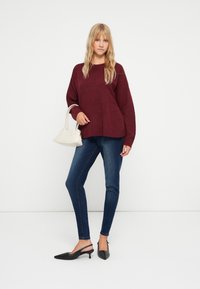 Bordeauxstickad tröja med lös passform, kombinerad med mörkblå slim-fit jeans. Tillbehören inkluderar en liten vit handväska och svarta spetsiga klackar.
