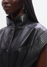 Zwarte leren mouwloze jas met hoge kraag, voorzien van drukknopen en gedetailleerd stikwerk op de schouders, die textuur en structuur benadrukken.