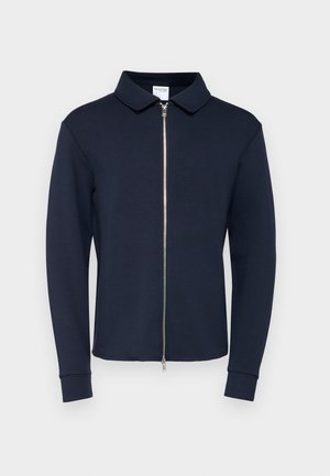 Tamno plava jakna na zip s ovratnikom, dugim rukavima i rebrastim manžetama, izrađena od glatkog, rastezljivog materijala. Srebrni detalj na zipu sprijeda.