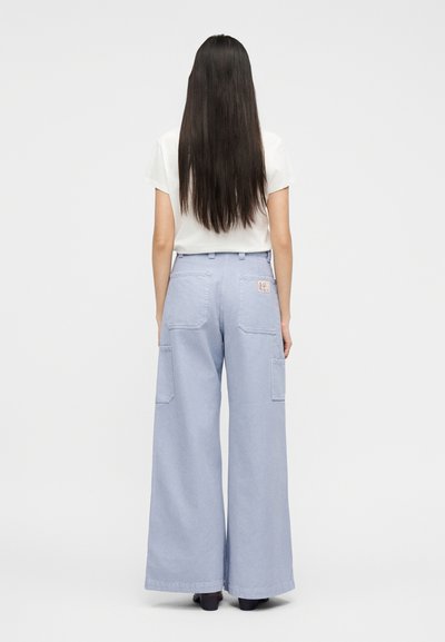 Γυναίκα με μακριά ίσια μαλλιά φορώντας λευκό t-shirt και ανοιχτό μπλε παντελόνι φαρδιάς γραμμής με τσέπες, στέκεται μπροστά σε απλό φόντο.