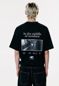 Una persona con capelli biondi corti indossa una maglietta nera con la scritta "In the middle of nowhere" e una foto di una strada in bianco e nero sul retro.