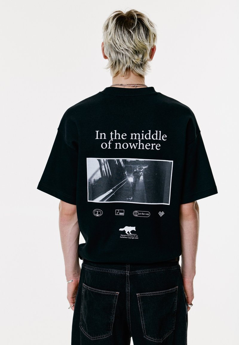 Persoon met kort blond haar draagt een zwart T-shirt met de tekst "In the middle of nowhere" en een zwart-wit foto van een weg op de achterkant.