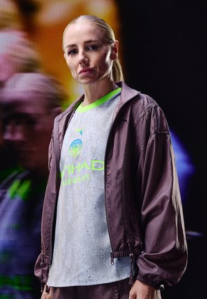 Femme aux cheveux blonds attachés porte un maillot de sport gris et vert fluo de Manchester City et une veste mauve, se tenant devant un fond sombre flou.