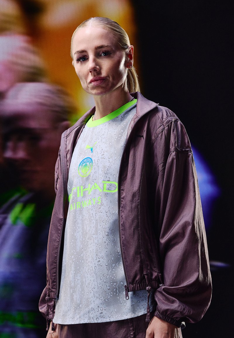 Moteris, turinti šviesius plaukus surištus atgal, vilki pilką ir neoninę žalią "Manchester City" sportinę marškinėlį ir purpurinę striukę, stovinti prieš neryškų tamsų foną.