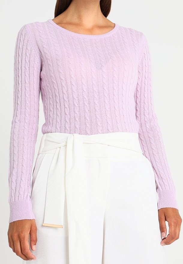 Femme portant un pull à manches longues en maille torsadée de couleur violet clair, rentré dans un pantalon blanc taille haute avec une ceinture nouée.