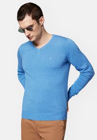 Niebieski sweter w serek z długimi rękawami, wykonany z miękkiego materiału. Posiada ribbowe mankiety i dół, z małym logo na klatce piersiowej.