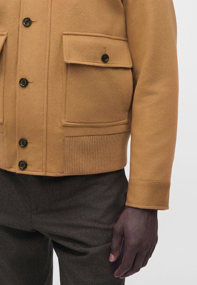 Homme portant une veste en laine camel marron boutonnée avec poches à rabat et pantalon sombre à carreaux sur fond uni.
