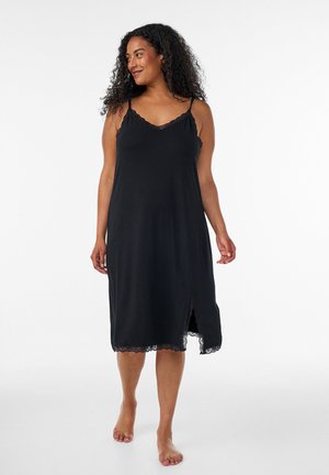 Femme aux cheveux bouclés portant une robe mi-longue noire sans manches avec une bordure en dentelle et une fente frontale, debout pieds nus sur un fond blanc.
