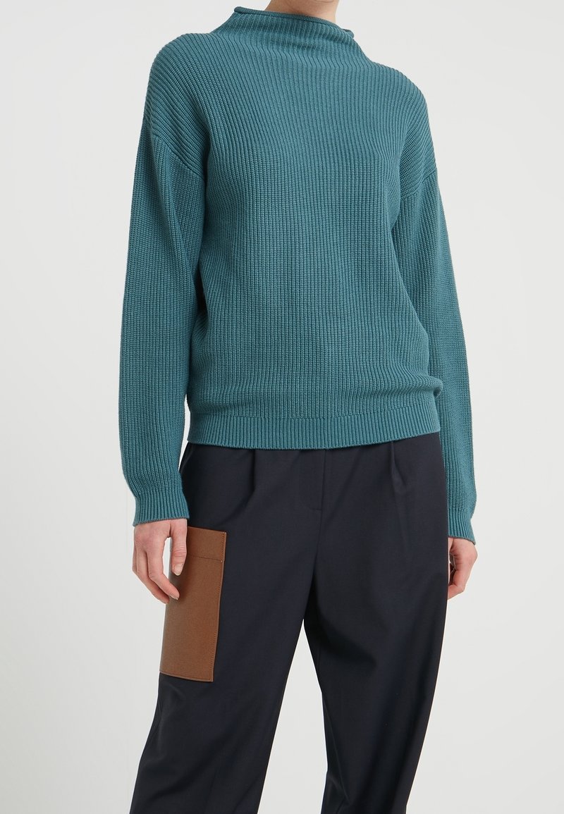 Maglione a coste in tonalità teal con scollatura alta, maniche lunghe e vestibilità rilassata, abbinato a pantaloni blu navy con una tasca a toppa marrone.