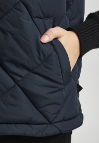 Navy quilted vest with a side pocket, featuring a textured fabric and a soft lining. The hem is stitched for durability.