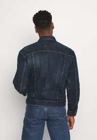 Dark blue denim jacket with a classic cut, prominent stitching, and a single patch at the upper back. Features snap buttons on cuffs.