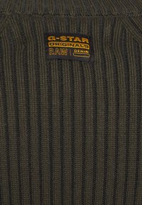 Maglione a coste verde scuro con etichetta G-Star Originals. Presenta un tessuto testurizzato e cuciture gialle a contrasto sull'etichetta.