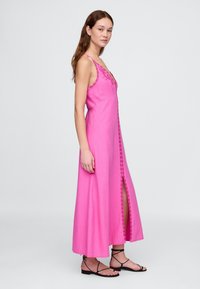 Rosa Maxikleid mit verstellbaren Spaghettiträgern, verziert mit bestickten Details und einem hohen Schlitz am Oberschenkel. Der Stoff hat eine glatte Textur.