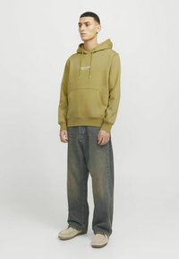Olivgrüne Kapuzenjacke mit Fronttasche, Kordelzügen und weißem Logo. Kombiniert mit lockeren dunkelblauen Jeans und beigen Schuhen.