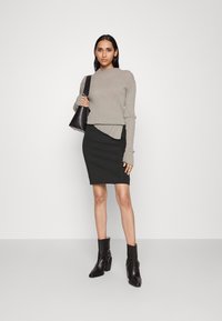 Hellgrauer, gerippter Pullover mit einem einzigartigen Saum, kombiniert mit einem schwarzen, taillierten Rock und schwarzen Stiefeletten mit markanten Beschlägen.