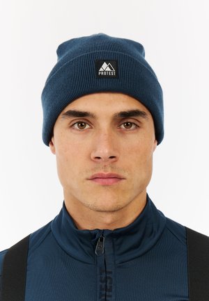 Dark blue knit beanie with ribbed texture and folded brim, featuring a black square patch with a mountain logo labeled "PROTEST."