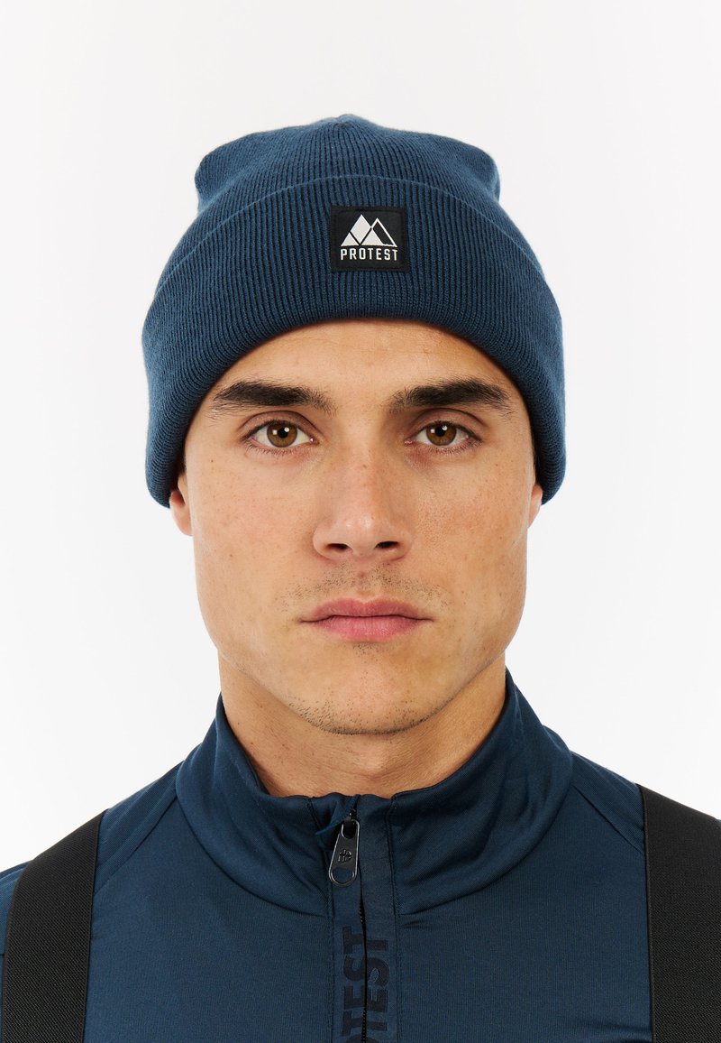 Dark blue knit beanie with ribbed texture and folded brim, featuring a black square patch with a mountain logo labeled "PROTEST."