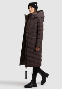 Langer brauner Daunenmantel mit Chevron-Nähten, hohem Kragen, Reißverschluss vorne und Seitentaschen. Eine schwarze Strickmütze und Ankle Boots runden den Look ab.