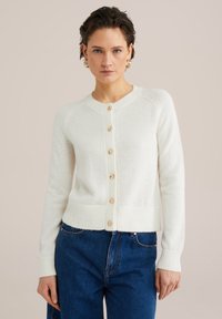 Vit stickad cardigan med rund halsringning, ribbade ärmslut och nederkant, samt sex träknappar framtill. Bärs över blå jeans.