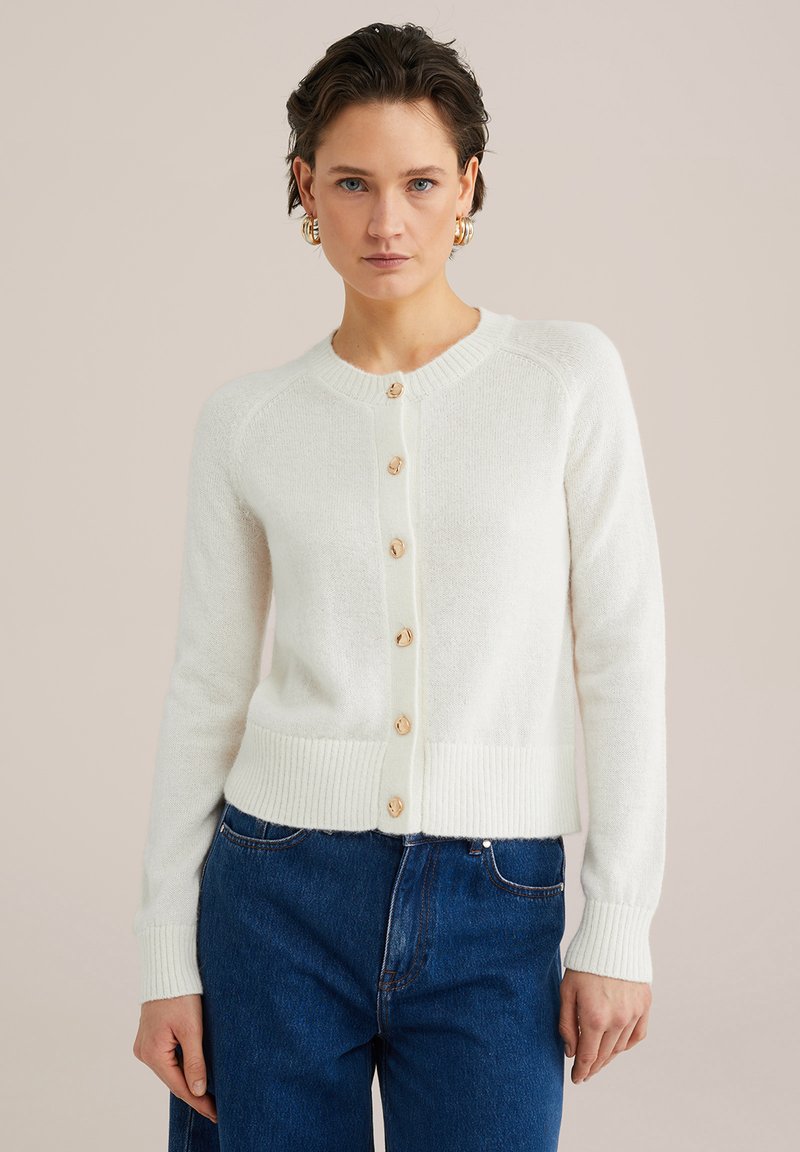 Vit stickad cardigan med rund halsringning, ribbade ärmslut och nederkant, samt sex träknappar framtill. Bärs över blå jeans.