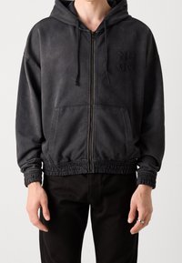Schwarze Zip-Hoodie mit strukturierter Oberfläche, ausgestattet mit einer Kapuze mit Kordelzug, elastischen Bündchen, Kängurutaschen und besticktem Logodesign.