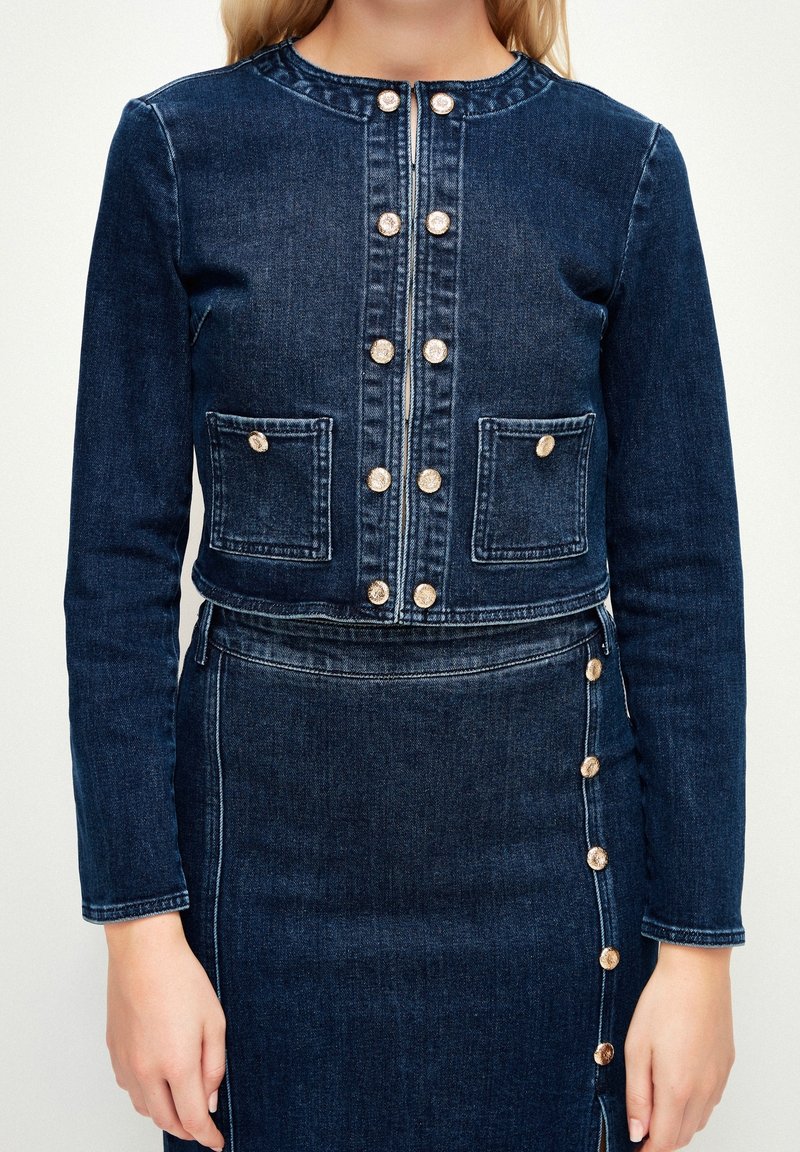 Femme portant une veste en denim bleu foncé avec des boutons dorés et une jupe taille haute assortie avec des détails de boutons dorés sur le côté.