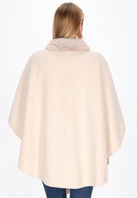 Poncho bej cu un guler moale din blană ecologică. Prezintă un design lejer, margini rotunjite și o textură netedă, purtat peste blugi din denim.