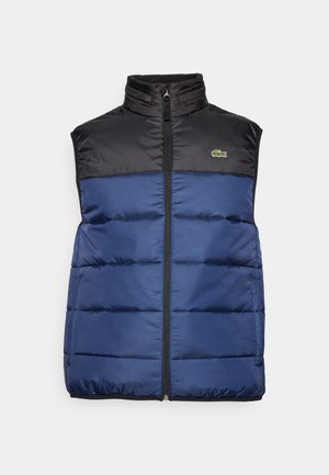 Gilet con imbottitura di colore blu navy, con una sezione superiore nera, colletto alto e zip frontale. Presenta cuciture a rombo orizzontali e un piccolo logo.