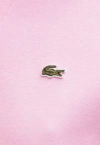 Πράσινο λογότυπο κροκόδειλου κεντημένο σε υφή απαλό ροζ ύφασμα, στο κέντρο της εικόνας.