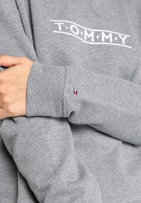 Grijze sweatshirt met "TOMMY" gedrukt op de borst en klein Tommy Hilfiger-vlaglogo geborduurd op de mouwmanchet.