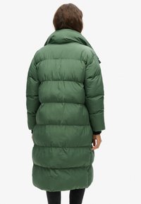 Giacca puffe verde con colletto alto, design trapuntato e silhouette lunga. Presenta una texture liscia e una finitura leggermente lucida.