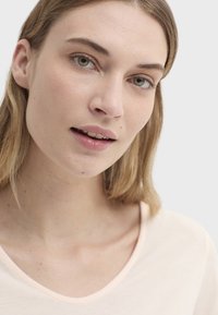 T-shirt rose pêche clair à col en V, en tissu doux et lisse. L'encolure présente des coutures contrastantes. Les traits du modèle sont neutres et subtils.