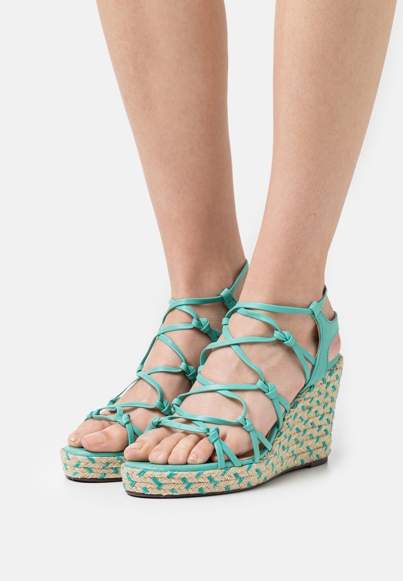 Sandales compensées turquoise avec plateforme en jute tressé, dotées de lanières croisées et de détails noués, mettant en valeur les orteils et les côtés texturés.