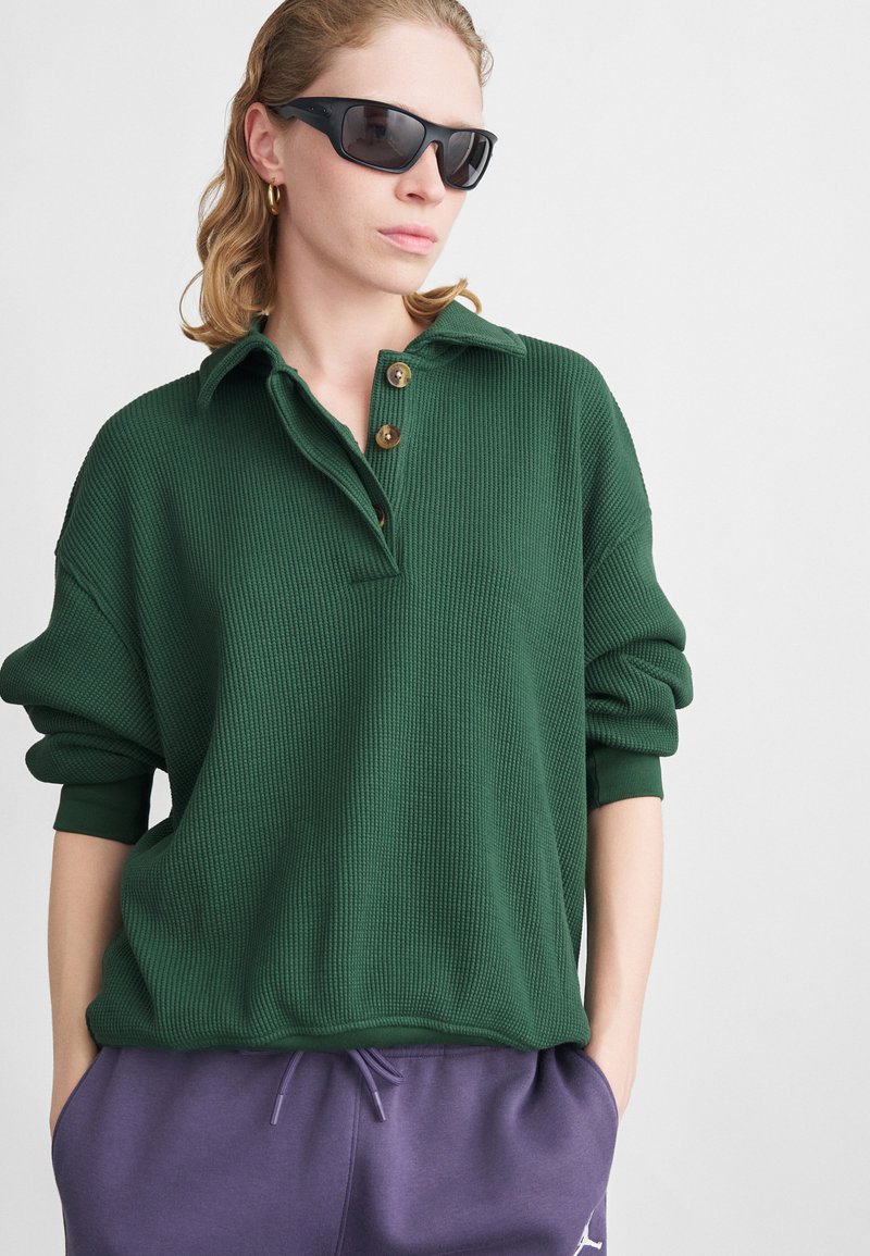 Suéter verde acanalado con cuello y tres botones, combinado con pantalones de chándal morados. La tela parece tener textura y un corte relajado. Gafas de sol negras.