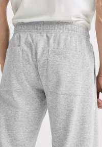 Man draagt lichtgrijze joggingbroek met elastische tailleband en twee grote achterzakken, draagt een wit shirt.