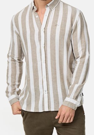 Homme portant une chemise à manches longues avec de larges rayures verticales beiges et blanches et un pantalon marron, debout avec les mains près des poches.