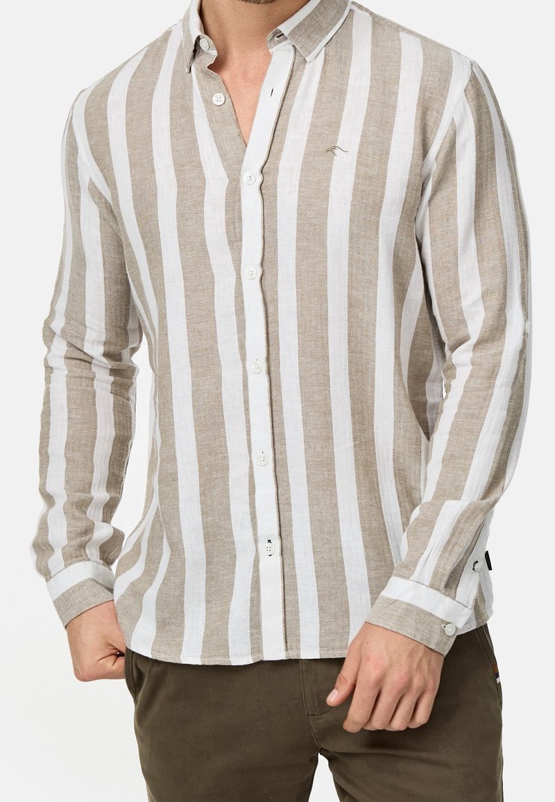 Homme portant une chemise à manches longues avec de larges rayures verticales beiges et blanches et un pantalon marron, debout avec les mains près des poches.