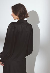 Black satin blouse with a collared neckline, puffed long sleeves, and a relaxed fit. Smooth texture with a soft sheen and slight drape.