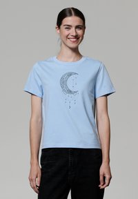T-shirt en coton bleu clair avec un graphique de croissant de lune noir et des accents suspendus. Manches courtes et design à col rond classique.