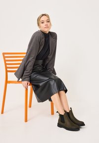 Blazer marrone sopra una maglia nera a coste, abbinato a una gonna in pelle nera e stivali alla caviglia verde oliva, seduto su una sedia arancione.
