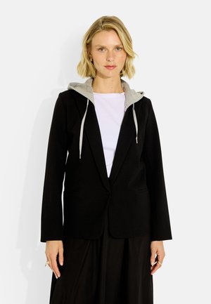 Mujer con pelo rubio corto que lleva una blazer negra sobre una camiseta blanca con capucha gris y pantalones negros sobre un fondo claro.
