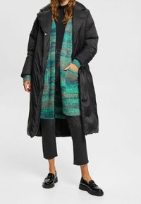 Czarna kurtka puchowa nałożona na zielony i niebieski sweter z dzianiny, połączona z czarnymi spodniami w krótkiej długości i błyszczącymi czarnymi mokasynami. Widoczne teksturowane wzory.