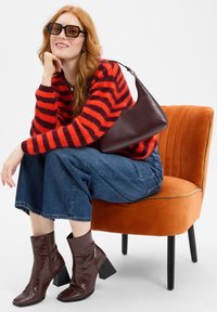 Rood en paars gestreepte fuzzy trui, blauwe wijde jeans, donkerbruine leren laarzen en een donkere handtas, zittend op een oranje fluwelen stoel.