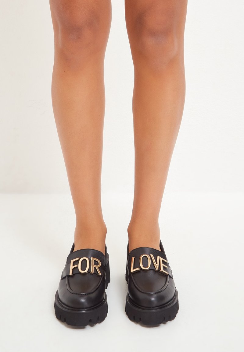 Jambes portant des mocassins noirs épais avec des lettres dorées formant "FOR" sur la chaussure gauche et "LOVE" sur la chaussure droite, sur un fond blanc uni.