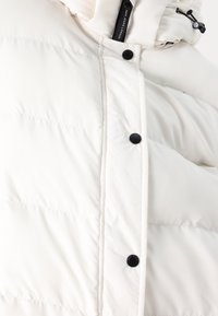 White puffer jacket with a quilted texture, black snap buttons, and a drawstring hood. Features a smooth, glossy exterior.